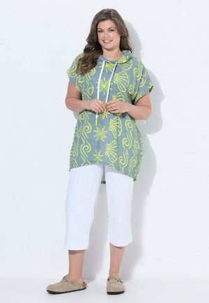 Vrouw in een groene capuchonjurk met gele zon- en schelpmotieven, witte capri-broek en beige instapschoenen, staand tegen een effen achtergrond.