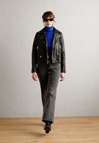 Black leather biker jacket over a blue turtleneck, paired with gray flared jeans and black ankle boots. Model wears dark sunglasses.