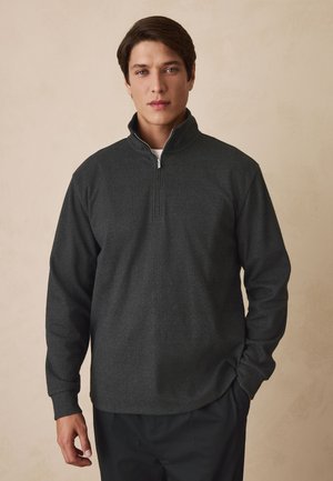 Ung mand, der vender fremad, iført en mørkegrå pullover med kvart lynlås og mørke bukser, stående mod en beige baggrund.