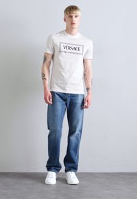 Tricou alb din bumbac cu logo-ul negru "VERSACE", blugi din denim albastru și adidași albi. Design simplu, croi standard, fără modele suplimentare.