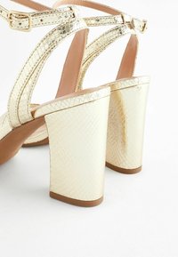 Guldiga högklackade sandaler med en texturerad yta, utrustade med breda klackar och stödjande vristremmar.