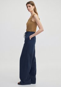 Tans mouwloos topje gecombineerd met marineblauwe wijde broeken met een trekkoord in de taille en zijzakken, een gladde textuur en contrasterende stiksels.
