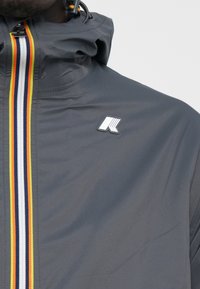 Hallideņiga hall jope, millel on lukuga avamine ja mitmevärviline triip (punane, kollane, tumesinine) ning logo päts. Sile, veekindel kangas.