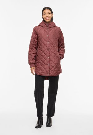 Woman wearing a maroon quilted hooded coat with snap buttons, black pants, and black ankle boots, standing against a white background.