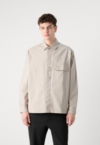 Light beige button-up shirt with a subtle texture, collared neckline, front pocket, and embroidered logo on the chest. Long sleeves.