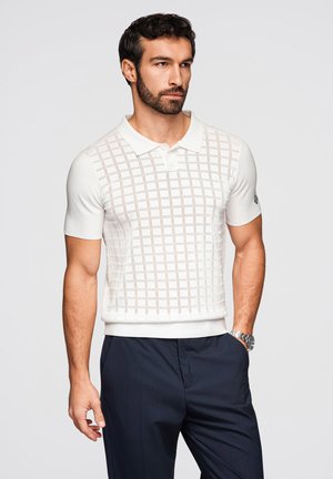 Uomo con capelli scuri e barba indossa una polo bianca a maniche corte con motivo a quadrati e pantaloni scuri, in piedi con la mano destra in tasca.