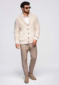 Beiger gestrickter Cardigan mit Schalkragen, kombiniert mit einem weißen Hemd, beigen Hosen und hellbraunen Wildlederstiefeln. Verfügt über Knöpfe und Taschen.