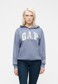 Νεαρή γυναίκα με ξανθά μαλλιά φοράει μπλε φούτερ GAP και σκούρο τζιν, στέκεται μπροστά σε απλό λευκό φόντο.