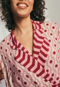 Raudonos ir rožinės spalvos audinys su geometriniais raštais; turi apykaklę su įpjovimu ir kontrastingas prisiuostytas juostas. Minkšta tekstūra, lengvas audinys.