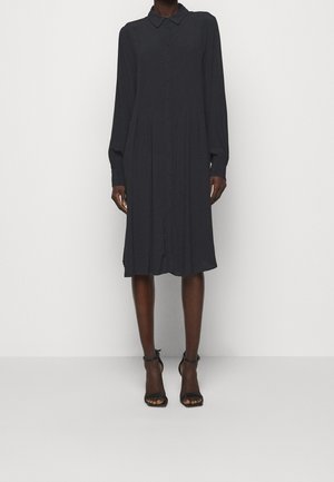 Personne portant une robe-chemise noire à manches longues, longueur genoux, avec col et boutonnage sur le devant, associée à des talons noirs à bride à la cheville.