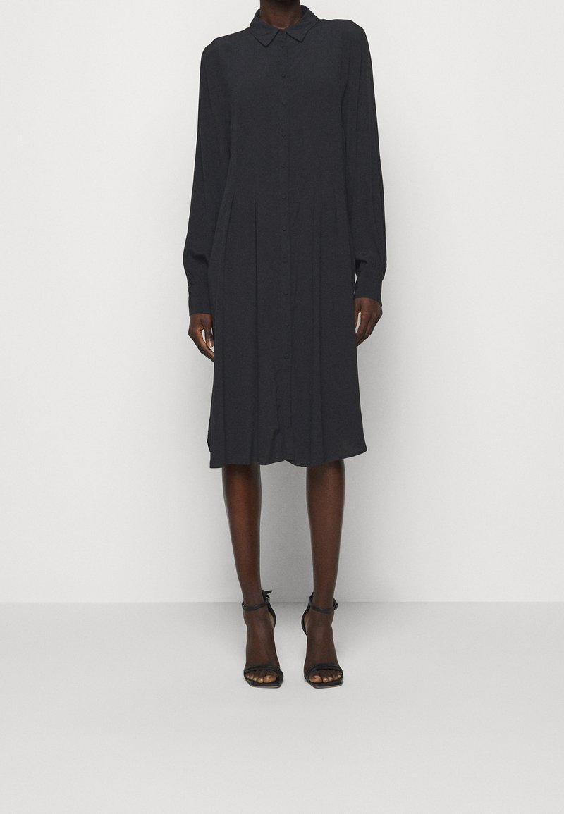 Personne portant une robe-chemise noire à manches longues, longueur genoux, avec col et boutonnage sur le devant, associée à des talons noirs à bride à la cheville.