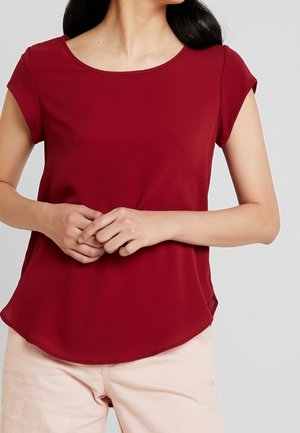 Femme aux longs cheveux noirs portant un chemisier rouge à manches courtes et un pantalon de couleur claire, mains jointes devant, regardant vers la droite.