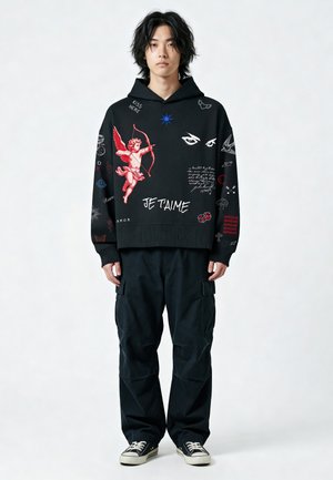 Giovane uomo con capelli neri che indossa una felpa con cappuccio nera con Cupido rosso e scritta bianca "JE T'AIME", pantaloni cargo neri e sneakers nere, in posizione neutra.