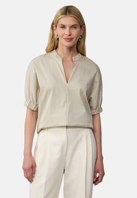 Hellbeige Bluse mit einem Latzkragen, kurzen Rüscheärmeln und einer lockeren Passform. Kombiniert mit cremefarbenen Hosen für einen abgestimmten Look.