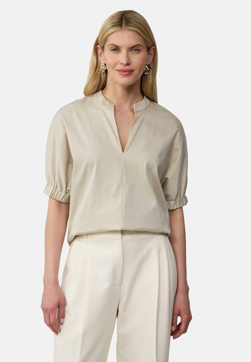 Hellbeige Bluse mit einem Latzkragen, kurzen Rüscheärmeln und einer lockeren Passform. Kombiniert mit cremefarbenen Hosen für einen abgestimmten Look.