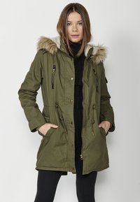 Olīvu zaļš parka ar mākslīgo kažokādu uz kapuces, ar rāvējslēdzēja kabatām, auklu vidukli un pogu aizdari; raksturīgs brīvā, relaksētā piegriezumā.