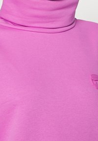 Top de cuello alto rosa hecho de una tela suave con un diseño ajustado. Presenta un sutil detalle de logo en la parte delantera para añadir interés.