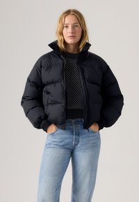 Chaqueta acolchada negra, con un cuello alto y cierre frontal de cremallera. Usada sobre un suéter estampado, combinada con unos jeans de tiro alto en azul claro.