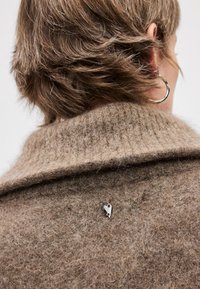 Cardigã de lã bege com uma gola alta e uma textura suave, apresentando um pequeno alfinete em forma de coração prateado preso na parte de trás.