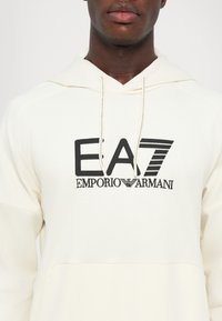 Felpa crema con cappuccio caratterizzata da un logo EA7 Emporio Armani ricamato in nero, cappuccio regolabile con stringhe, tessuto morbido e tasca frontale.