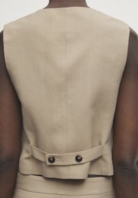 Vista posteriore di una persona che indossa un gilet senza maniche beige con un dettaglio di cintura buttonato sulla parte bassa della schiena, abbinato a pantaloni coordinati.