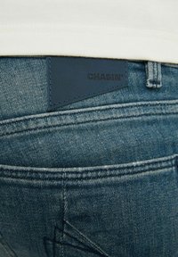 Džínové kalhoty s modrým odstínem, s koženou visačkou označenou "CHASIN'," stehováním na kapsách a texturovaným povrchem.