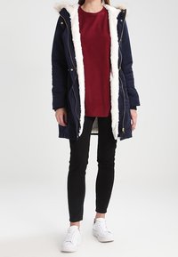 Marineblauer Parka mit Kunstfellfutter und Kapuze, kombiniert mit einem roten Pullover, schwarzen Skinny-Hosen und weißen Sneakers. Verfügt über Reißverschlüsse und Kordeln.