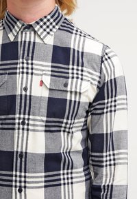 Marineblauw en witte geruite blouse met een kraag, voorzien van twee voorkruiszakken en een contrasterend geruit patroon. Zachte stofstructuur.