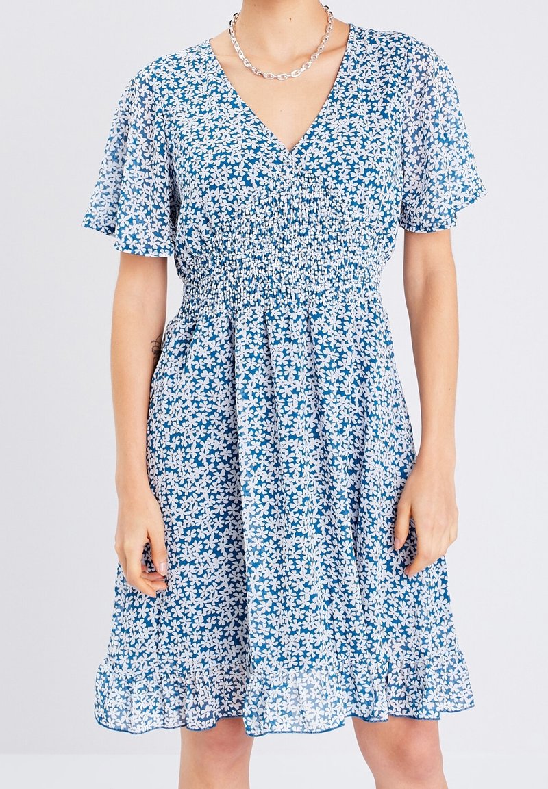 Robe bleue à motif floral blanc, manches courtes volantées, encolure en V, taille smockée et ourlet à volants, portée avec un collier chaîne en argent.