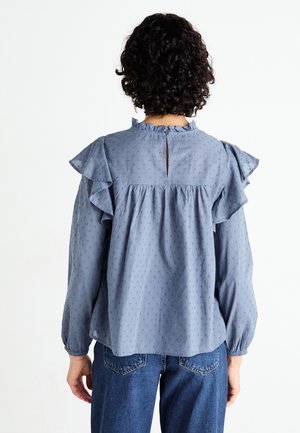 Osoba o kręconych, ciemnych włosach, ubrana w jasnoniebieską, długorękawową bluzkę z falbankami na ramionach i małym kluczem z tyłu, w połączeniu z niebieskimi dżinsami.