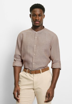 Mann trägt ein hellbraunes Hemd mit Mandarin-Kragen und aufgekrempelten Ärmeln sowie beige Hosen mit braunem Gürtel, steht vor einem weißen Hintergrund.