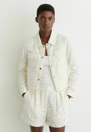 Femeie cu păr scurt, purtând o jachetă albă din dantelă cu imprimeu floral și pantaloni scurți asortați, stând cu mâinile în buzunare, pe un fundal alb.