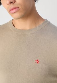 Beige gestrickter Pullover mit Rundhalsausschnitt und kleinem roten gestickten Logo auf der Brust, das eine glatte, weiche Textur zeigt.