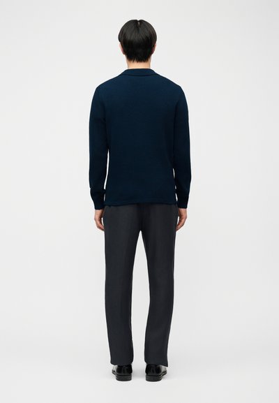 Homme debout de dos, portant une chemise à manches longues bleu foncé, un pantalon noir et des chaussures noires, sur un fond clair uni.