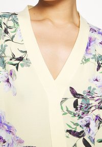 Floral blouse in light yellow fabric. Features a V-neck design with purple and green floral patterns. Smooth texture with ribbed accents.