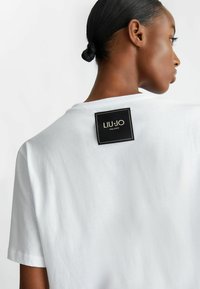 T-shirt bianco in cotone con maniche corte e scollatura a girocollo, con un'etichetta del marchio nera e dorata sul retro. Tessuto liscio.