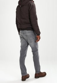 Mørk polstret jakke med hætte, grå jeans med baglommer og brune støvler, set bagfra mod en hvid baggrund.