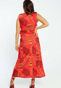Robe maxi rouge avec imprimé feuilles tropicales en orange et marron. Style sans manches avec une coupe ample et un détail de lien sur le côté.