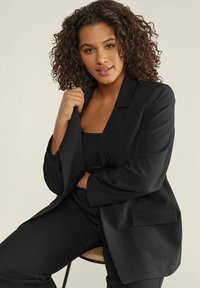 Blazer nero con un design su misura, caratterizzato da un colletto a punte e tasca frontale, indossato sopra una maglia nera e abbinato a pantaloni neri.