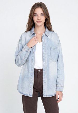 Jeune femme aux longs cheveux portant une veste en jean bleu clair sur un haut blanc et un pantalon marron foncé, debout devant un fond uni.