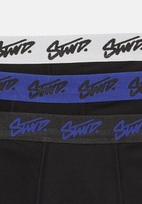 Svarta boxershorts med elastiska midjeband i vitt, blått och svart, vardera med varumärket "Stand." tryckt i fet, stiliserad text.