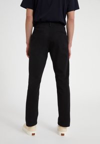 Person som bär svarta raka byxor, svart skjorta och beige sneakers, står mot en enkel vit bakgrund, bakifrån.