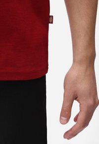 Camiseta de algodón roja con dobladillo y abertura lateral, combinada con pantalones de tela negros y una vista parcial de una mano descansando cerca de la ropa.