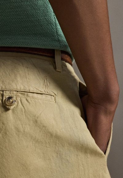 Osoba ubrana w beżowe spodnie z guzikiem przy tylnej kieszeni oraz zieloną teksturowaną koszulę wpuszczoną do środka, z ręką włożoną do kieszeni.