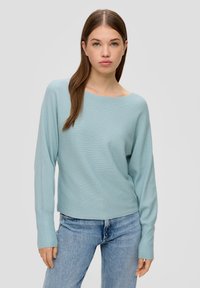 Hellblauer, gerippter Pullover mit weitem Rundhalsausschnitt und langen Ärmeln, kombiniert mit hellblauen Jeans. Die Textur wirkt weich.