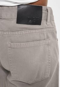 Jeans de mezclilla beige con una etiqueta de cuero negra en la cintura que dice "DEF", un bolsillo trasero y detalles de costura sutiles.