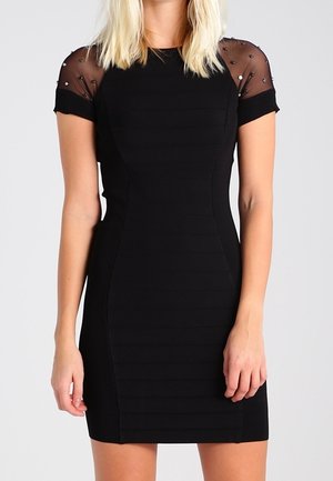 Frau trägt eng anliegendes schwarzes Kleid mit kurzen transparenten Ärmeln, verziert mit kleinen Perlen vor einfarbigem Hintergrund.