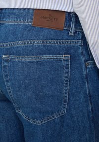 Jeans de mezclilla azul oscuro con una textura suave, que cuentan con una etiqueta de cuero marrón etiquetada "HACKETT" en la cinturilla y un detalle en el bolsillo trasero.