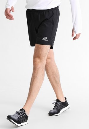 Mężczyzna idący, ubrany w czarne szorty Adidas, czarne i szare buty do biegania oraz białą bluzę z długim rękawem z otworami na kciuki.