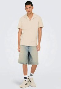 Camisa de manga curta bege com colarinho em bico, combinada com calções de ganga azul claro. O modelo usa ténis brancos e meias às riscas.